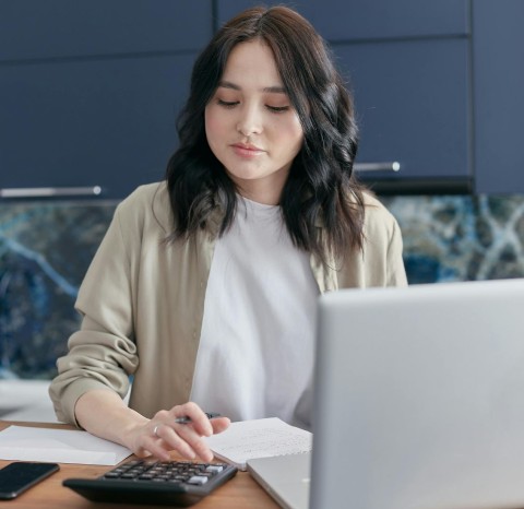 A woman with dark hair in a cardigan using a calculator sitting at a desk with a laptop